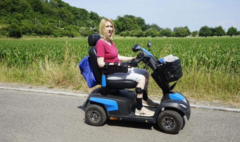 Personne touchée par une lésion cérébrale Une femme aux cheveux blonds est assise dans un fauteuil roulant électrique devant un champ à la végétation dense. L'expression de son visage est grave.