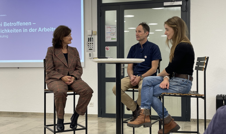 Drei Personen sitzen an einem hohen Tisch und sind im Gespräch. Im Hintergrund läuft eine Powerpoint-Präsentation.