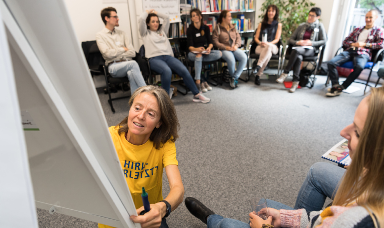 Eine Frau im gelben FRAGILE-T-Shirt schreibt auf ein Flipchart. Im Hintergrund sitzen mehrere Personen in einem Halbkreis und schauen zu.
